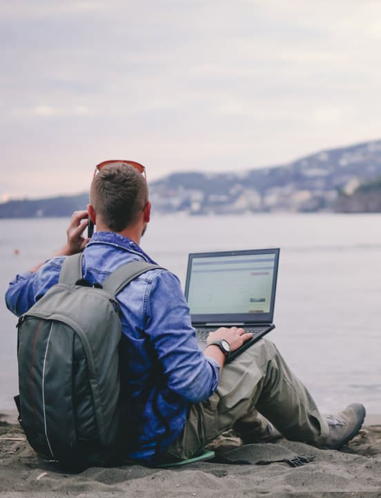 man sitting on lakeshore looking at laptop talking on phone