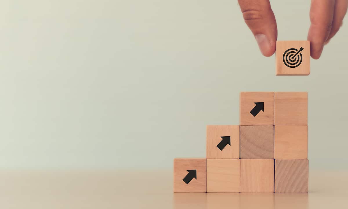 Hand puts the wooden cubes with "Goal" icon on the top of stacking wooden cubes. The goal is the transition of VLSC to Microsoft Admin Center for License Management.